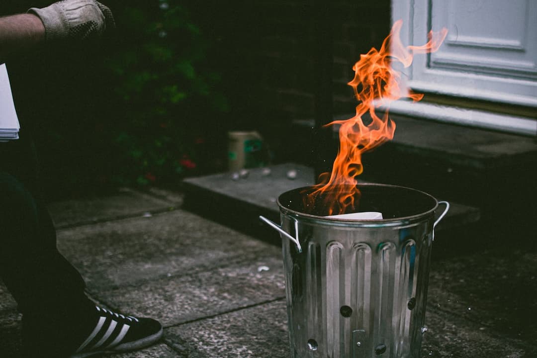 Metal trash bin with visible flames burning paper inside, person nearby wearing a glove and black Adidas sneakers, outdoor concrete surface near a door