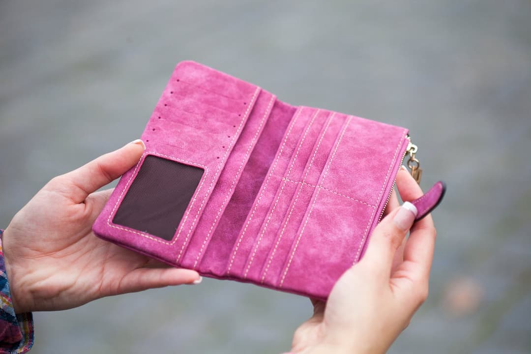 Empty pink suede wallet, held open by two hands, featuring card slots, ID window, and zipper pouch, with blurred gray pavement background