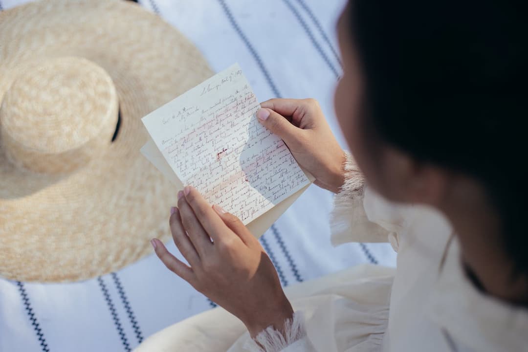 Woman in light clothing holding and reading a handwritten letter, seated on a white striped blanket, straw hat beside her