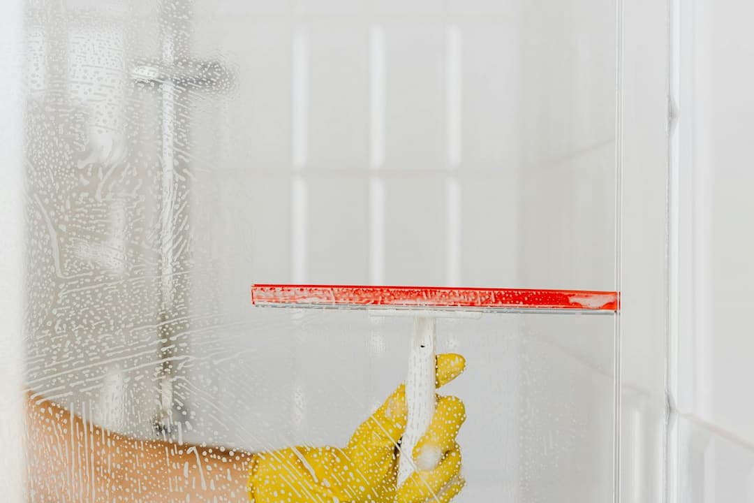 Person wearing yellow gloves using a red squeegee to clean soap suds off a glass shower door