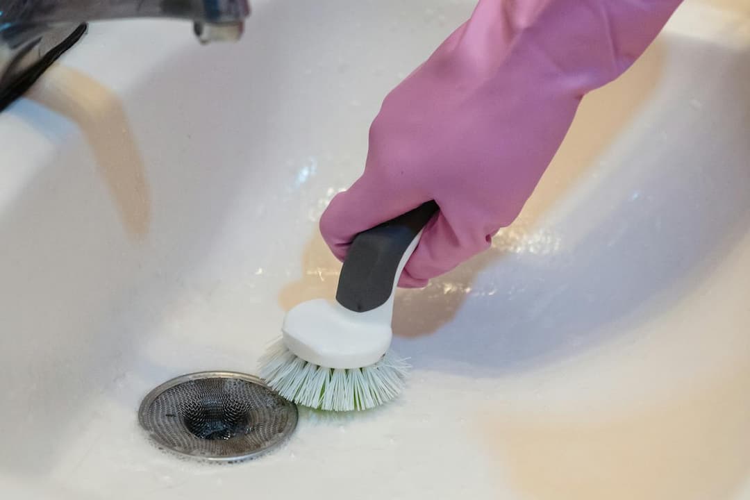 Pink gloved hand scrubbing white bathroom sink near metal drain with a round bristle cleaning brush