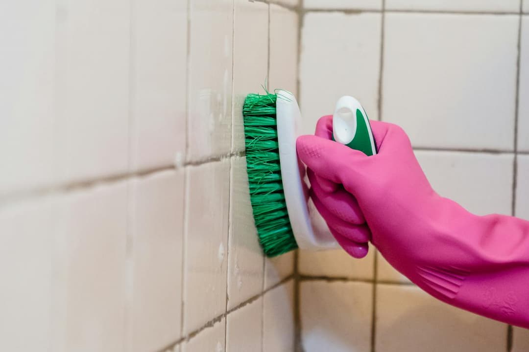 Hand in pink rubber glove scrubbing tiled wall with green-bristled cleaning brush