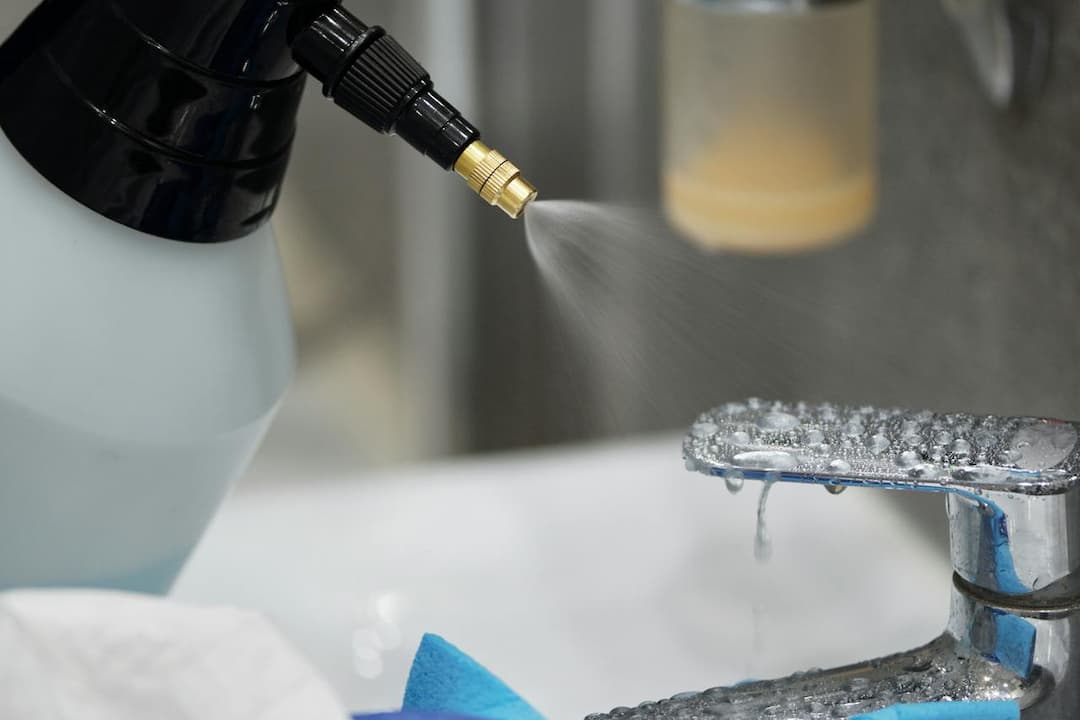 Spray bottle emitting fine mist onto a wet bathroom faucet, with cleaning cloths nearby