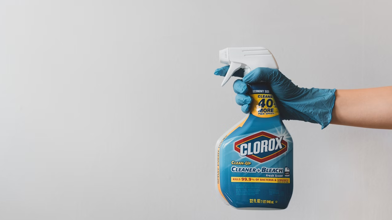 Gloved hand in blue nitrile glove, holding a white spray bottle, spraying clear bleach mist, against a tiled surface