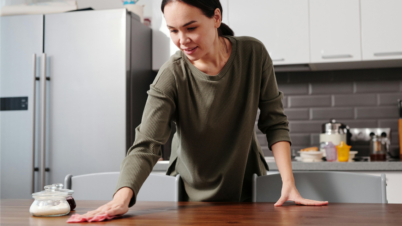 Woman wiping a wooden kitchen table with a cloth, modern kitchen background, fridge and countertop items visible