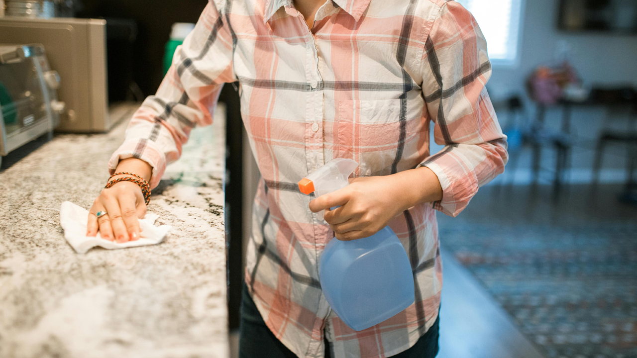woman wiping kitchen counter with cloth, standing near sink, cleaning supplies on surface, light streaming in from window, modern kitchen interior, focused posture