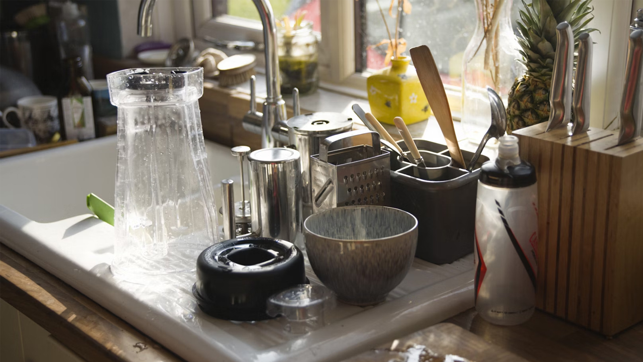Cluttered kitchen sink and countertop with unwashed dishes, blender parts, bowls, utensils, water bottle, knife block, and natural light streaming in through a window