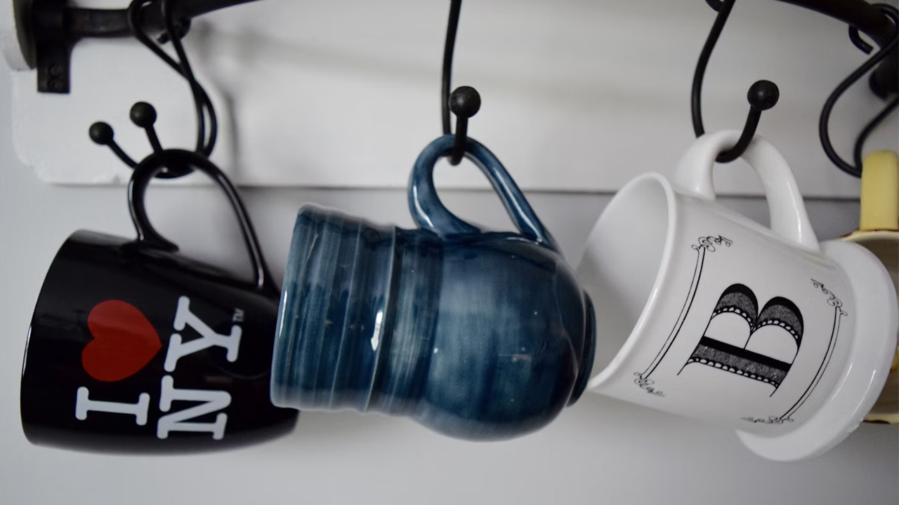 Three coffee mugs hanging on black hooks, including a black "I ❤️ NY" mug, a blue ceramic mug, and a white mug with the letter "B"