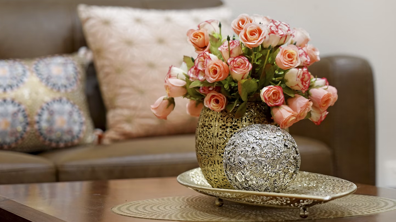 Coffee table centerpiece with pink roses in a gold perforated vase, paired with a decorative silver orb on a matching gold tray, brown sofa in background