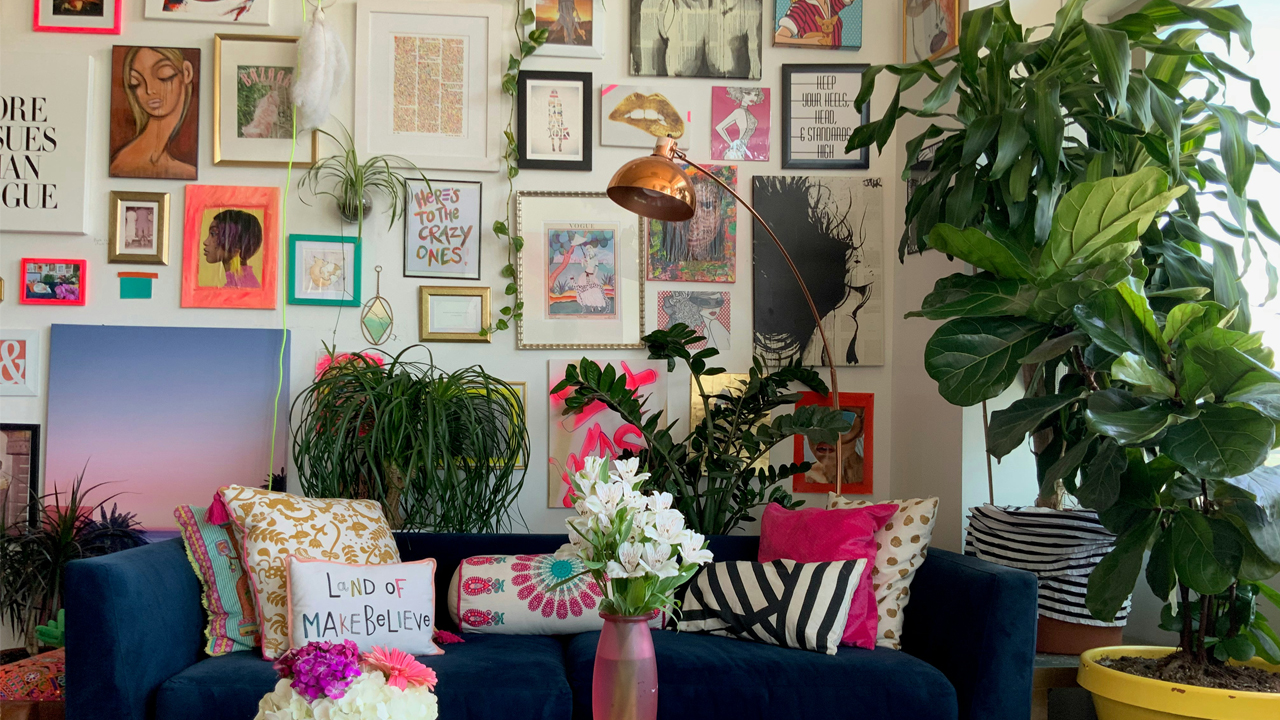 Colorful living room with navy blue couch, vibrant throw pillows, pink flowers in a vase, green houseplants, and a gallery wall filled with framed art and quotes