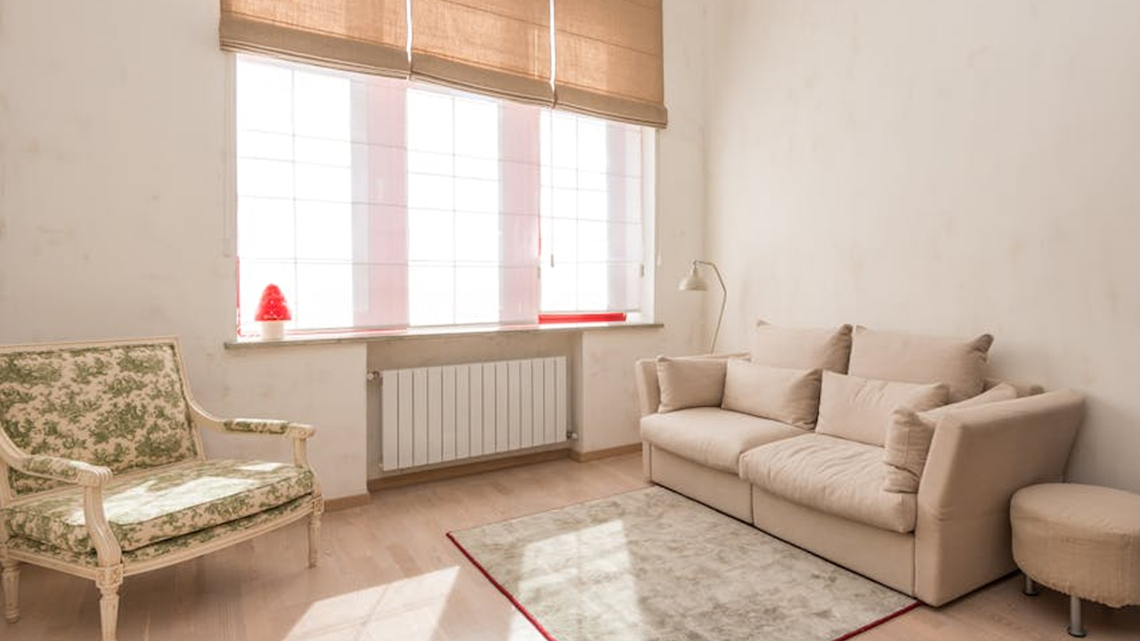 Minimalist living room with cream loveseat, floral-patterned armchair, small round stool, light area rug, tall window with beige blinds, red lamp on windowsill
