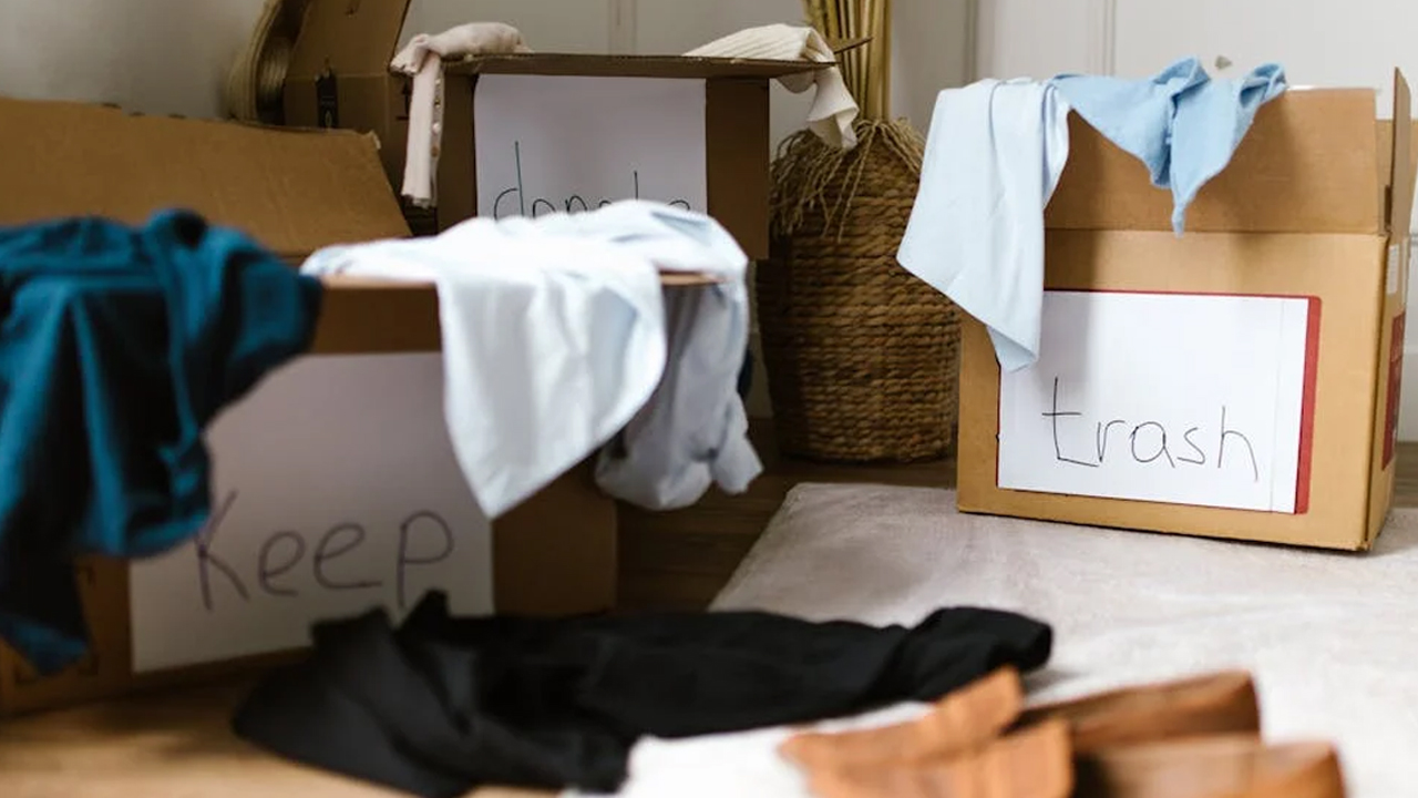 Cardboard boxes labeled "Keep," "Donate," and "Trash," overflowing with assorted clothes, placed on a floor during a decluttering session