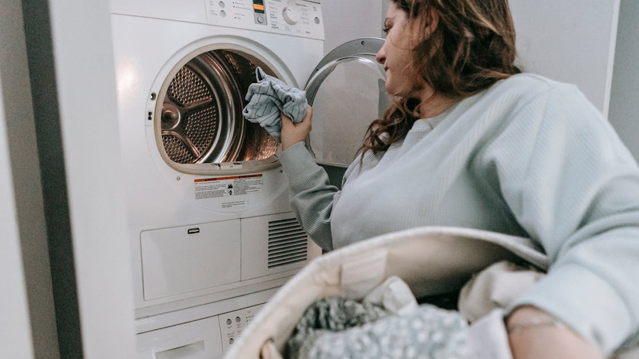 Woman loading clothes into a front-load dryer, holding laundry basket, wearing light grey sweatshirt