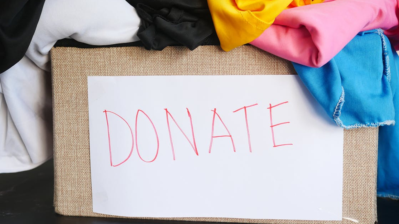 Cardboard donation box filled with colorful folded clothes, white paper label with red handwritten text "DONATE" taped to the front