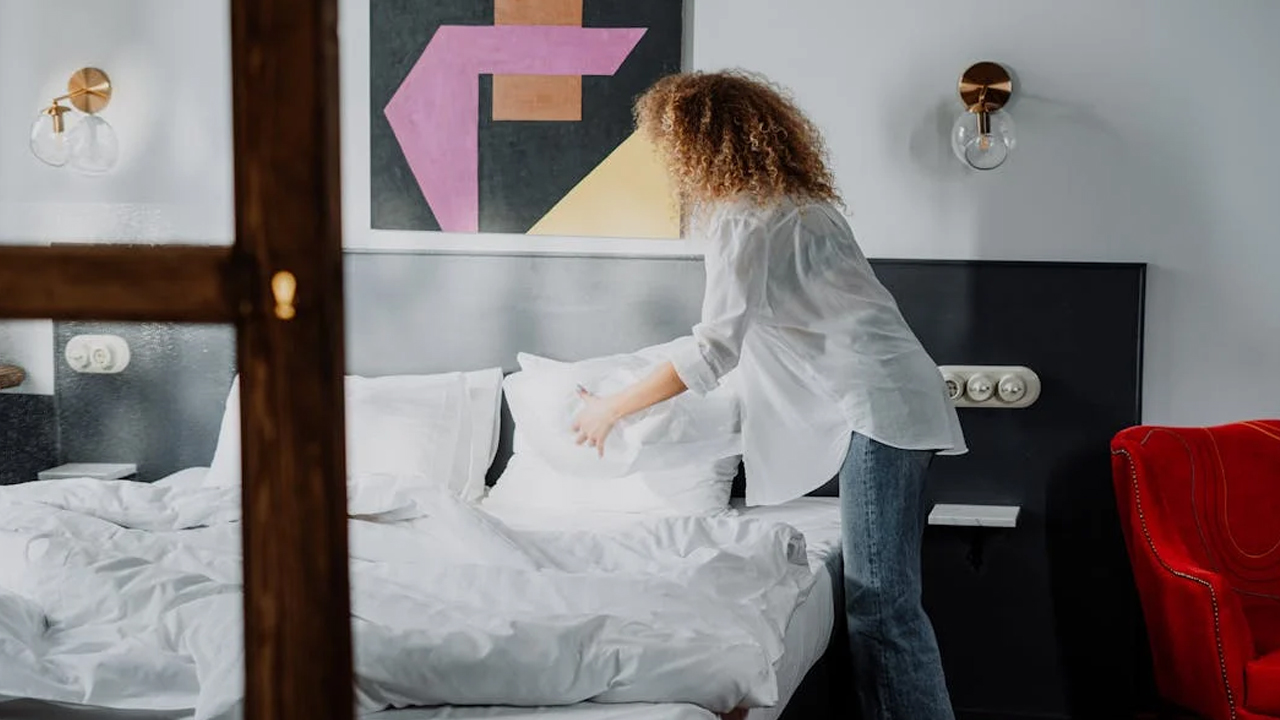 Woman with curly hair making a bed, white bedding, modern wall art, black headboard, wall-mounted lights
