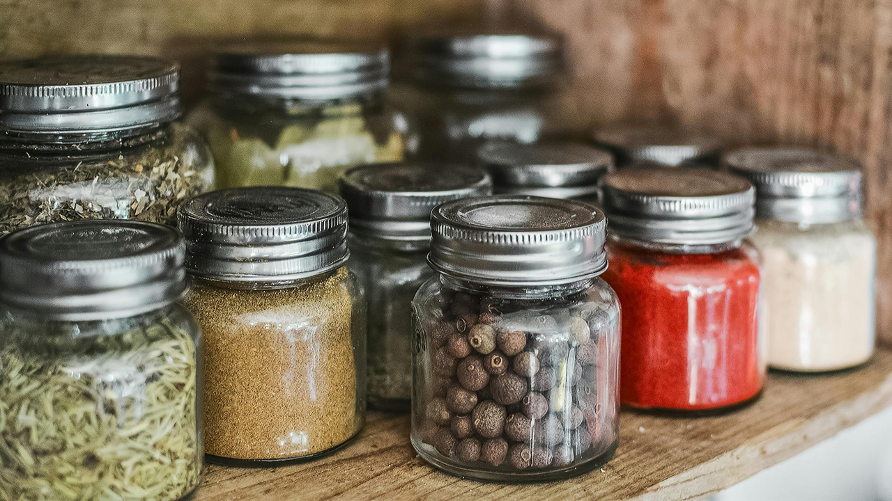 Assorted glass spice jars with metal lids arranged on a wooden shelf, filled with herbs, powders, and whole spices in various colors