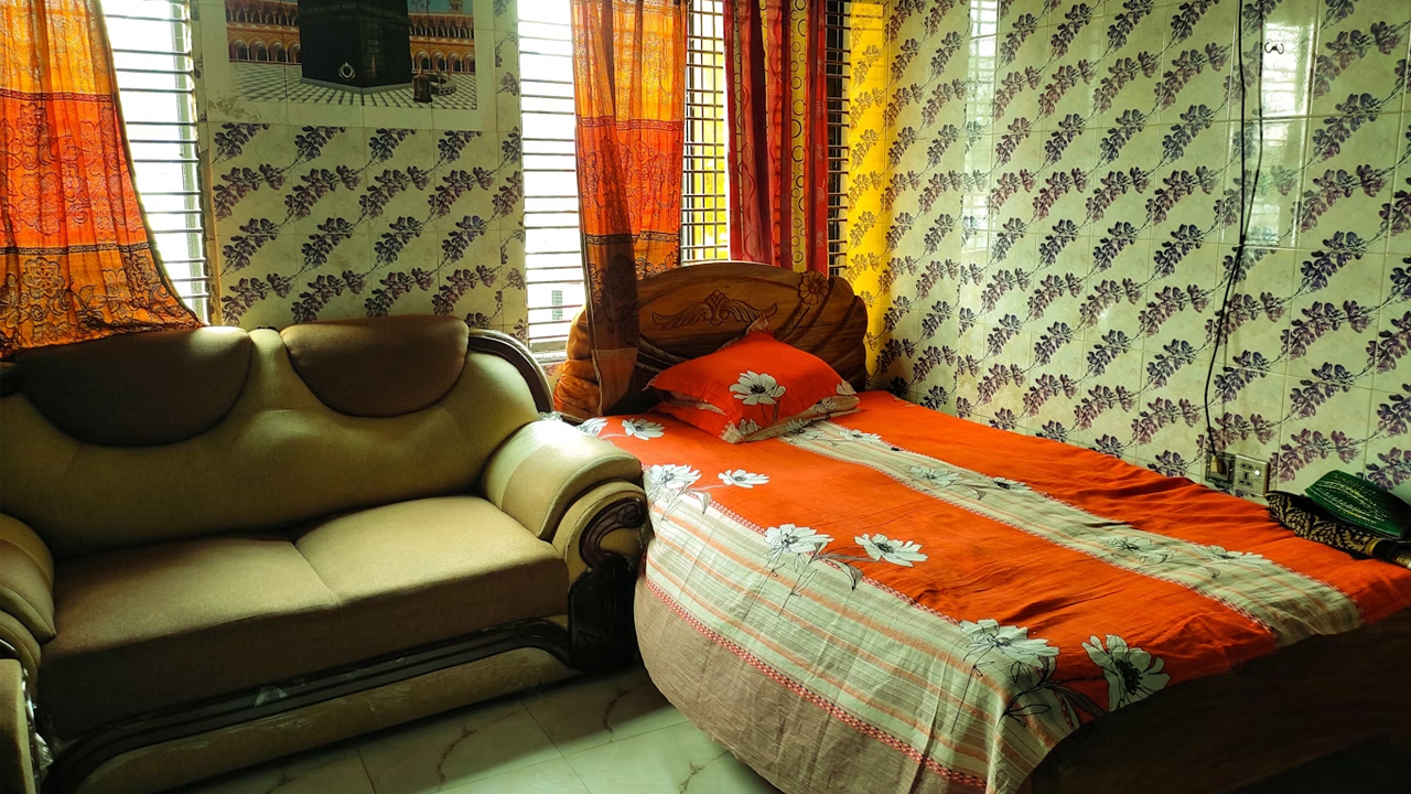 Small bedroom with a patterned wallpaper, wooden bed with orange floral bedding, beige sofa, and colorful curtains covering the windows