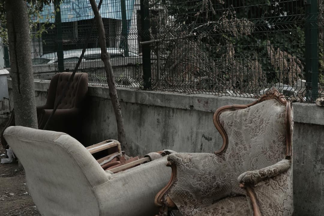 Abandoned vintage armchairs and a sofa discarded near a concrete wall and wire fence, surrounded by dry leaves and urban decay