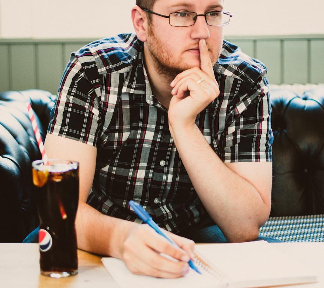Man in plaid shirt sitting on a couch, holding a pen and touching his face in thought, notebook open, glass of soda with straw on the table