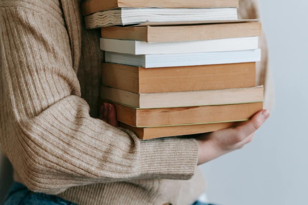 Person in beige sweater holding a tall stack of books, close-up, paperbacks with varied covers, plain background