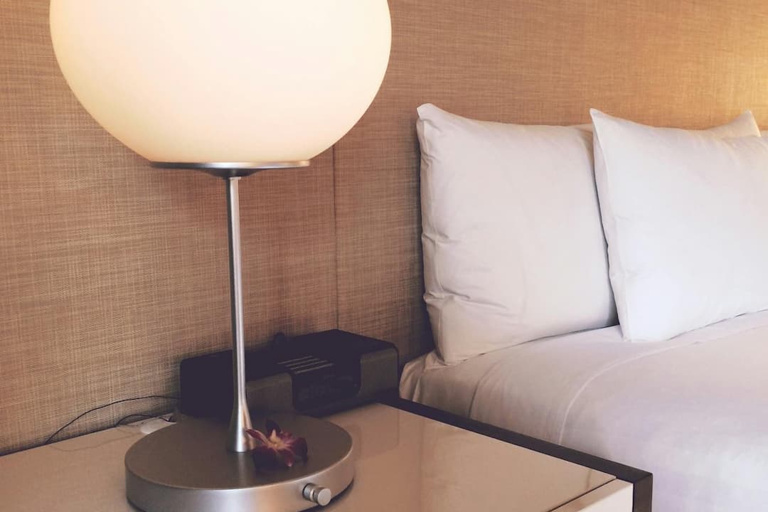 Modern nightstand with a round frosted glass lamp, a small flower on the lamp base, and a neatly made bed with white pillows in a softly lit bedroom