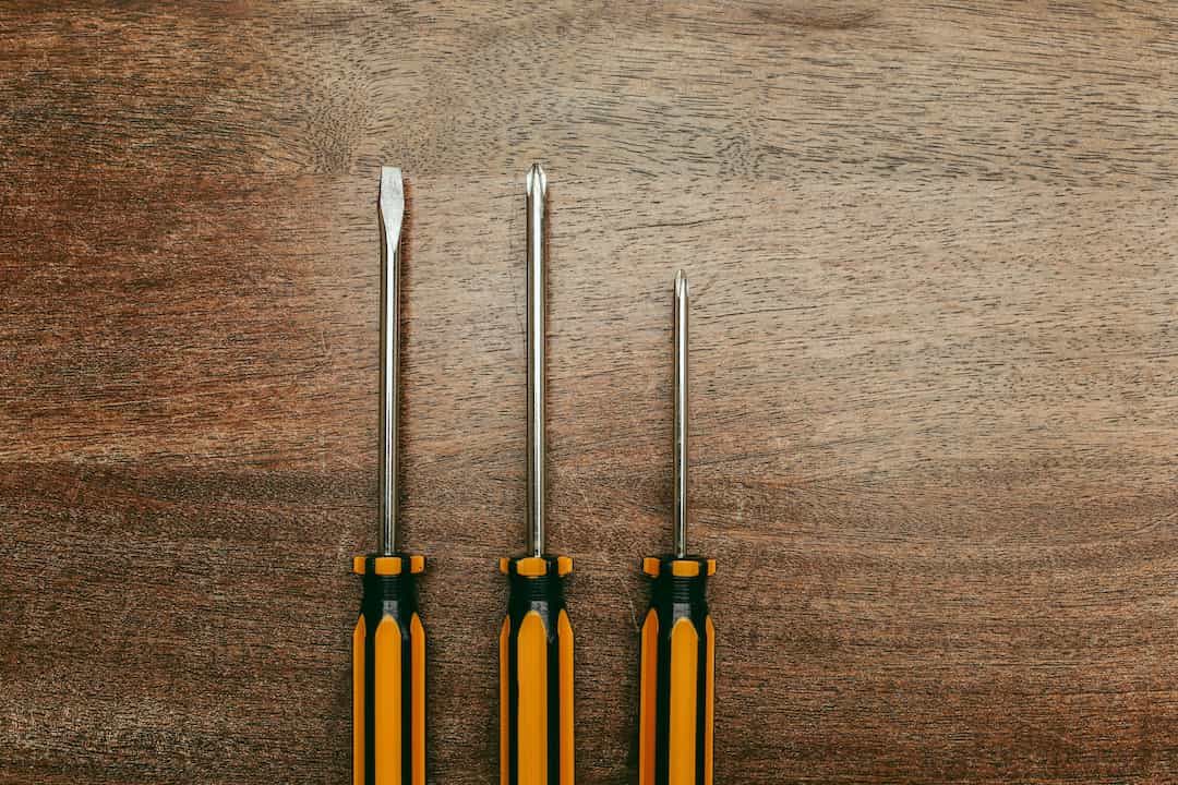 Three screwdrivers with yellow and black handles arranged vertically on a wooden surface; one flathead and two Phillips head screwdrivers