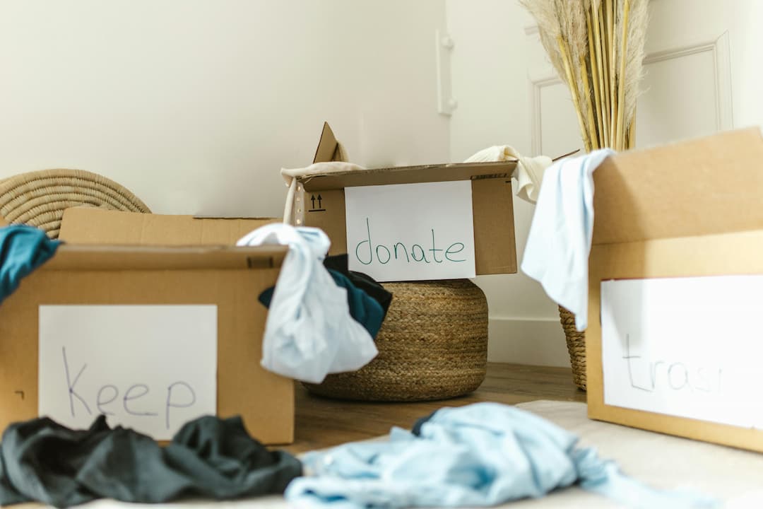 Three cardboard boxes labeled "keep," "donate," and "trash," filled with assorted clothing items, placed in a bright room with a wicker basket and pampas grass decor