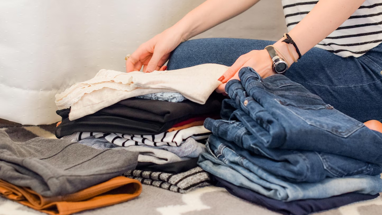 Person sorting and folding clothes on the floor, organized piles of jeans, shirts, and other garments, striped shirt and watch visible on their wrist