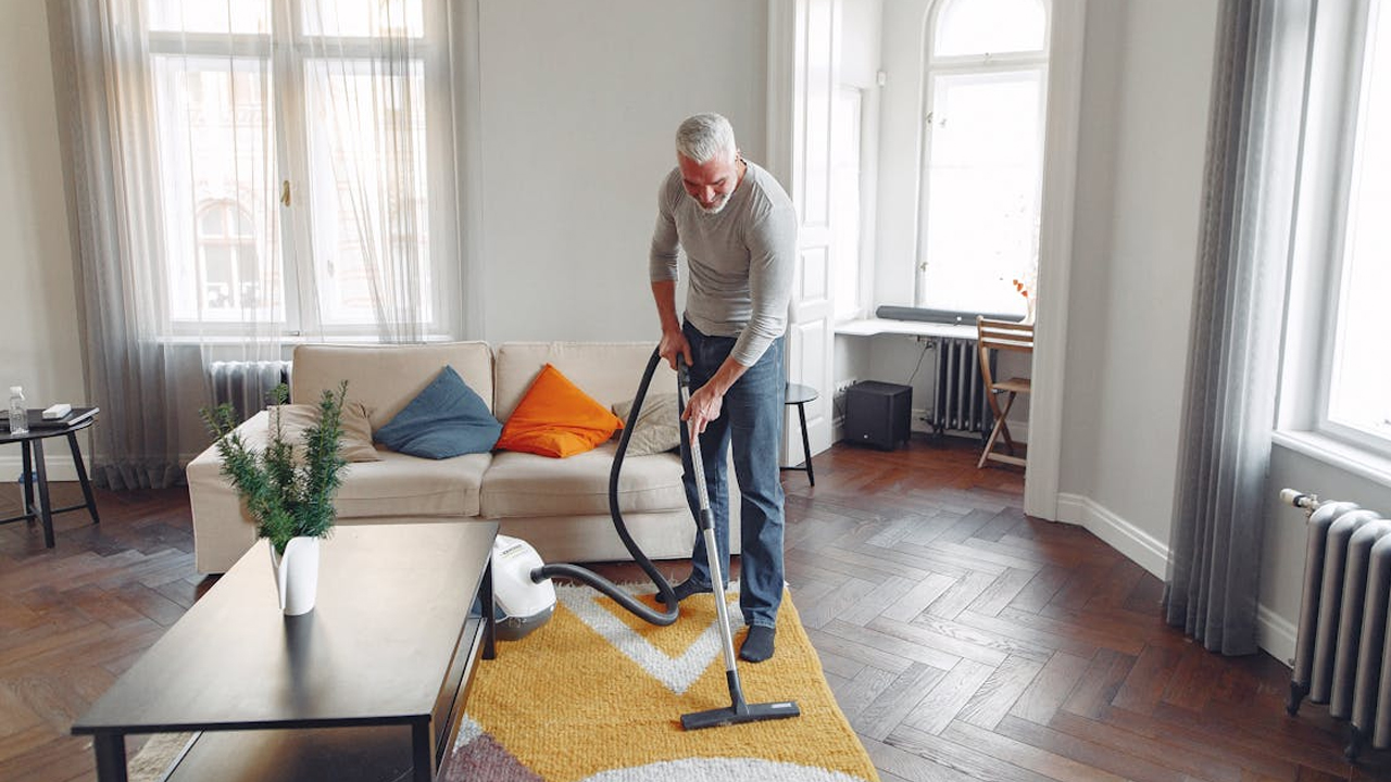 Older man vacuuming a patterned rug in a bright living room, modern decor, white walls, wood flooring