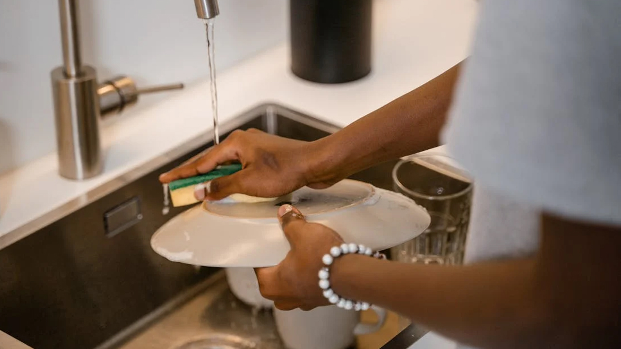 Person washing a white plate under running tap water with a green sponge, standing at a stainless steel kitchen sink