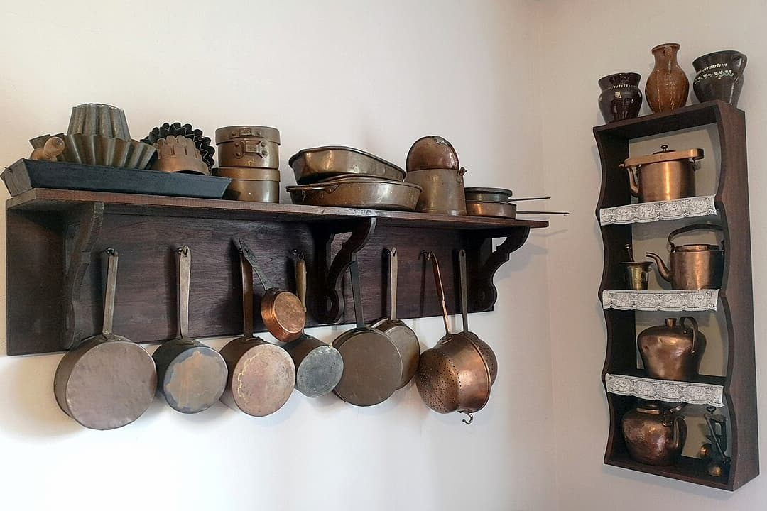 Wall-mounted wooden shelves displaying a collection of vintage copper cookware including saucepans, baking molds, kettles, and pitchers. Pans are hanging by their handles, and additional pots are stacked on the upper shelf and in a narrow side shelf with lace-trimmed edges