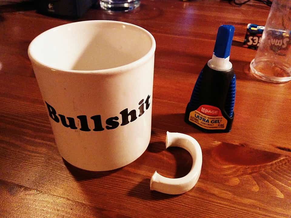 A white mug with the word "Bullshit" printed on it sits on a wooden table, next to its broken handle and a bottle of LePage Ultra Gel super glue