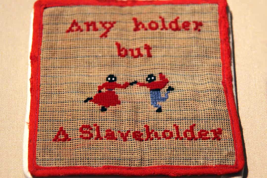 A vintage embroidered pot holder with a red border and the stitched slogan "Any holder but A Slaveholder." The center features two small figures, one in red and one in blue, holding hands and appearing to dance