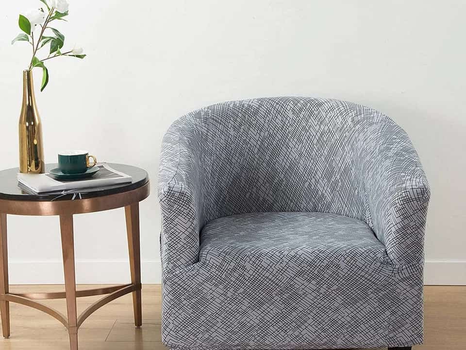 Gray U-shaped accent chair, geometric patterned upholstery, placed beside a round side table with black top and gold legs, green teacup and gold vase with white flowers on table, light wooden floor and white wall background