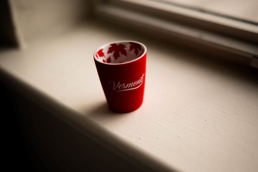 A small red cup with the word "Vermont" written on the front sits on a windowsill; the interior of the cup is decorated with red maple leaves