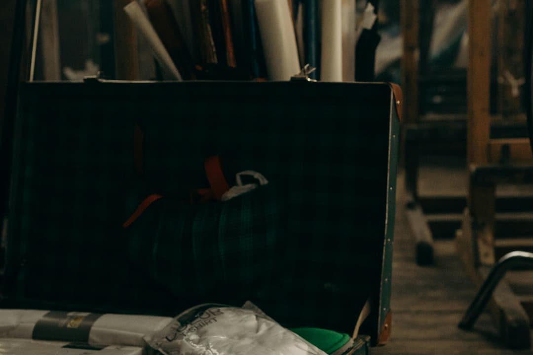 Dimly lit attic with an open suitcase, rolls of paper or fabric, and miscellaneous stored items, set against wooden rafters and floorboards