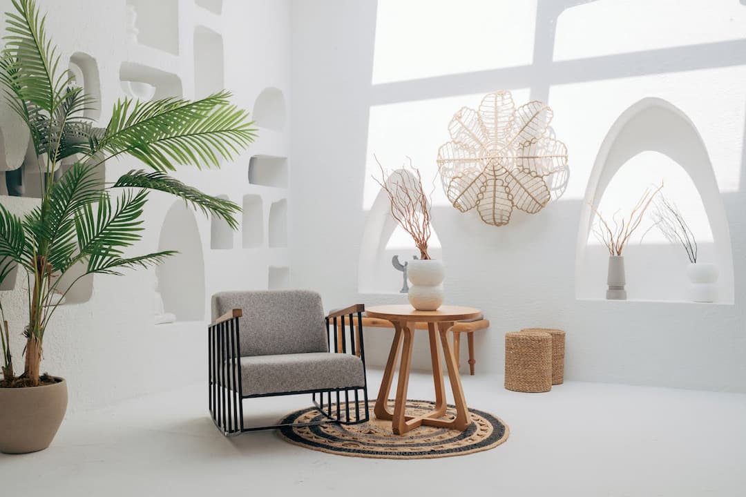 minimalist boho living space, white arched wall niches, grey armchair with black metal frame, round wooden table, decorative vases with twigs, woven rug, indoor palm plant, natural light casting shadows