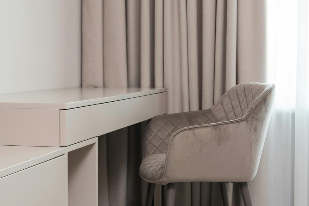 Modern beige chair with quilted backrest, positioned next to a sleek, minimalist beige desk, in front of floor-length beige curtains