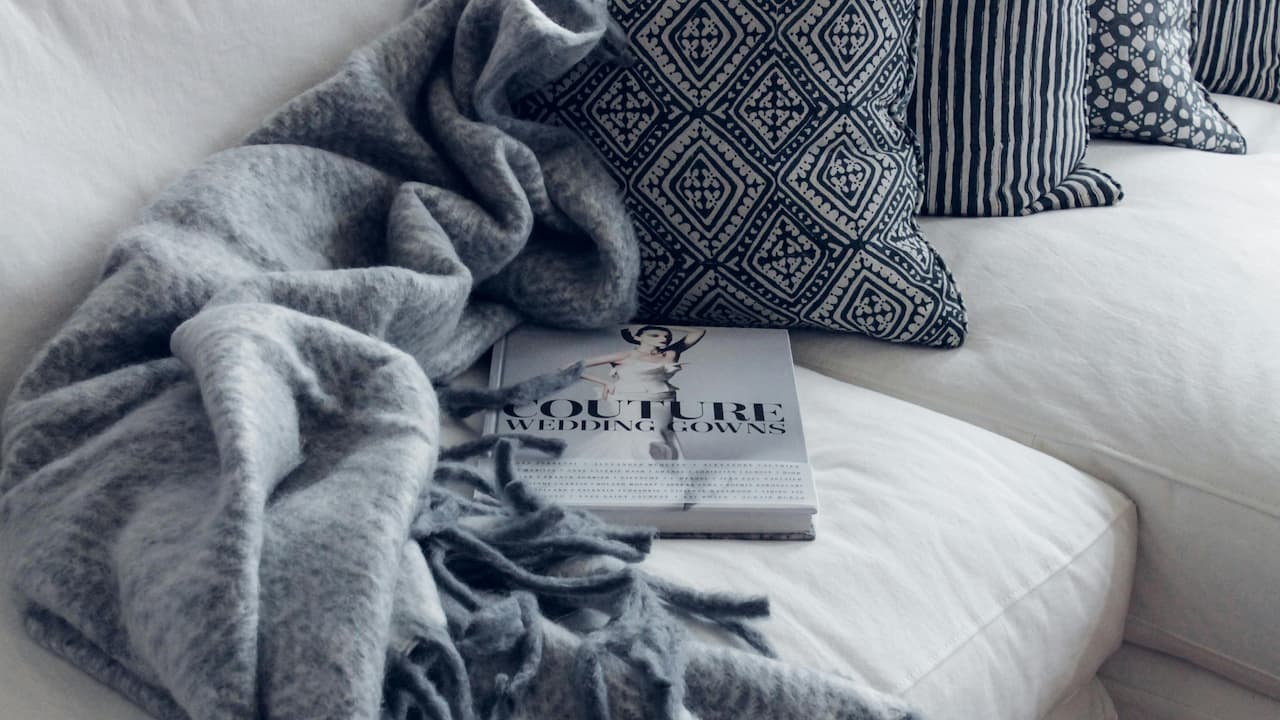 White couch, patterned navy throw pillows, grey wool blanket with tassels, fashion book titled "Couture Wedding Gowns" partially covered