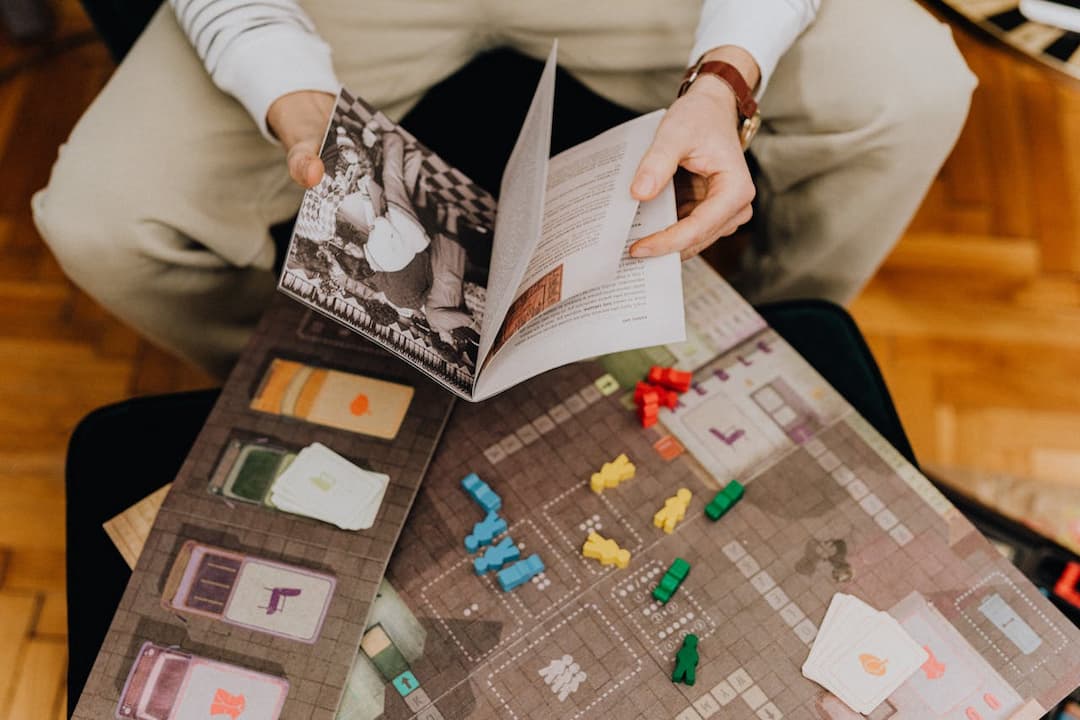 Person holding a game manual over a colorful board game with small red, yellow, green, and blue player tokens, and various cards spread out