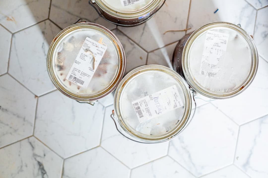 Four labeled paint cans with transparent lids, placed on white hexagonal tiled flooring with marble patterns