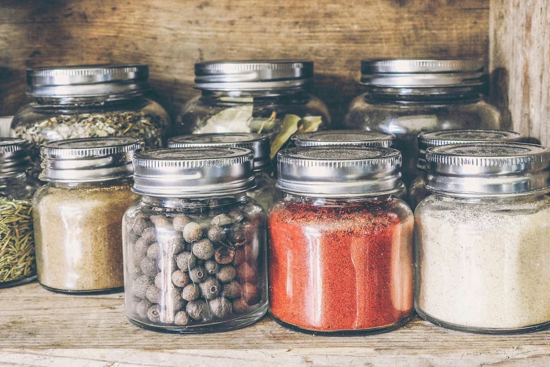 Glass jars filled with various dried herbs and spices, including black peppercorns, paprika, and bay leaves, arranged on a rustic wooden shelf