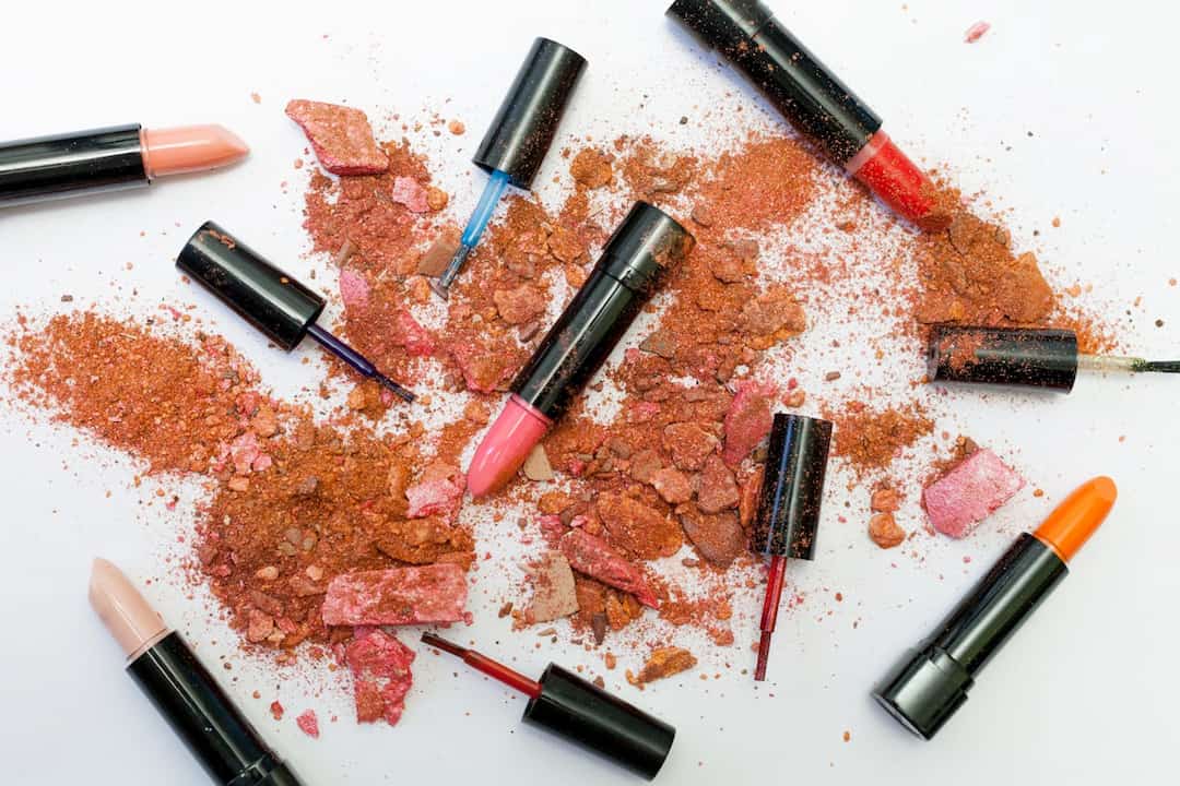 Assorted lipsticks and nail polish tubes scattered on a white surface, surrounded by crushed orange and pink blush or eyeshadow powders