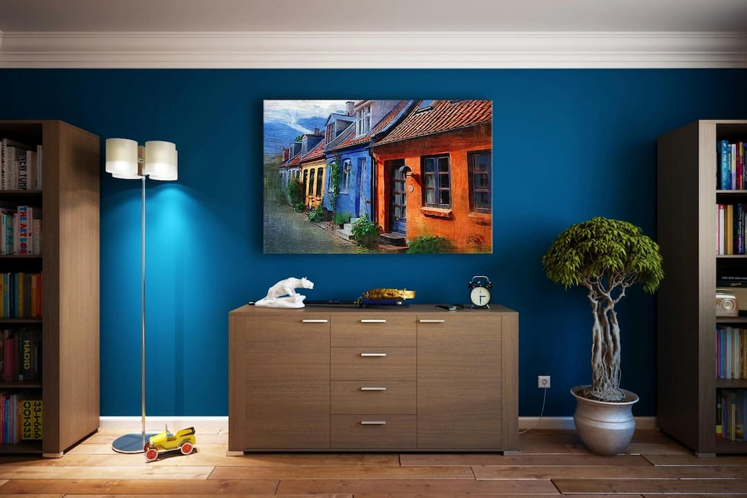 Modern living room with blue accent wall, wooden sideboard, colorful framed painting of houses, floor lamp, potted bonsai tree, bookshelves, and toy airplane on wooden floor