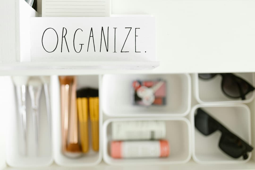 Organized white drawer with labeled compartments holding makeup brushes, lip balm, sunglasses, and a note reading