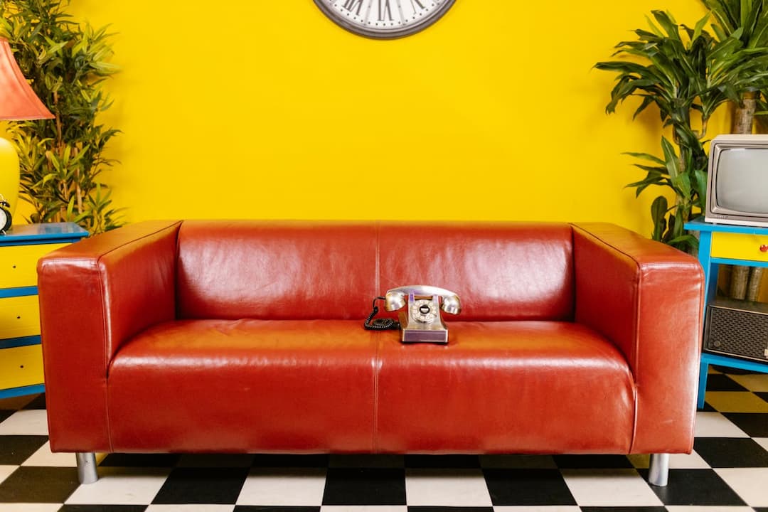 red leather sofa, rotary dial telephone on seat, yellow wall, black and white checkered floor, vintage TV on blue and yellow cabinet, decorative plants, wall clock with Roman numerals