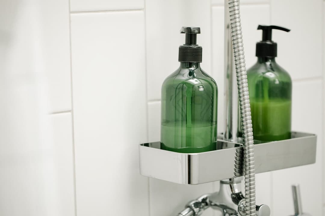 Green liquid soap bottles with black pump tops placed in a stainless steel shower caddy mounted on a tiled white bathroom wal