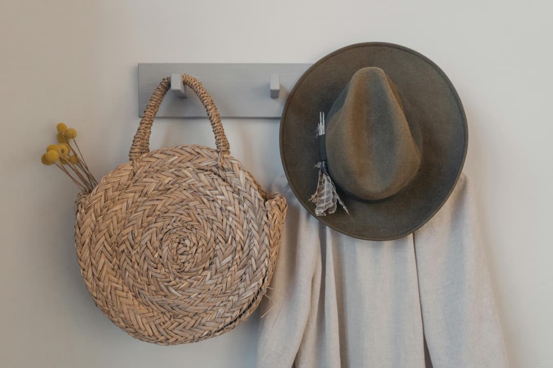 Woven round straw bag with yellow flowers, olive green fedora hat with feather, beige fabric hanging on wall hooks