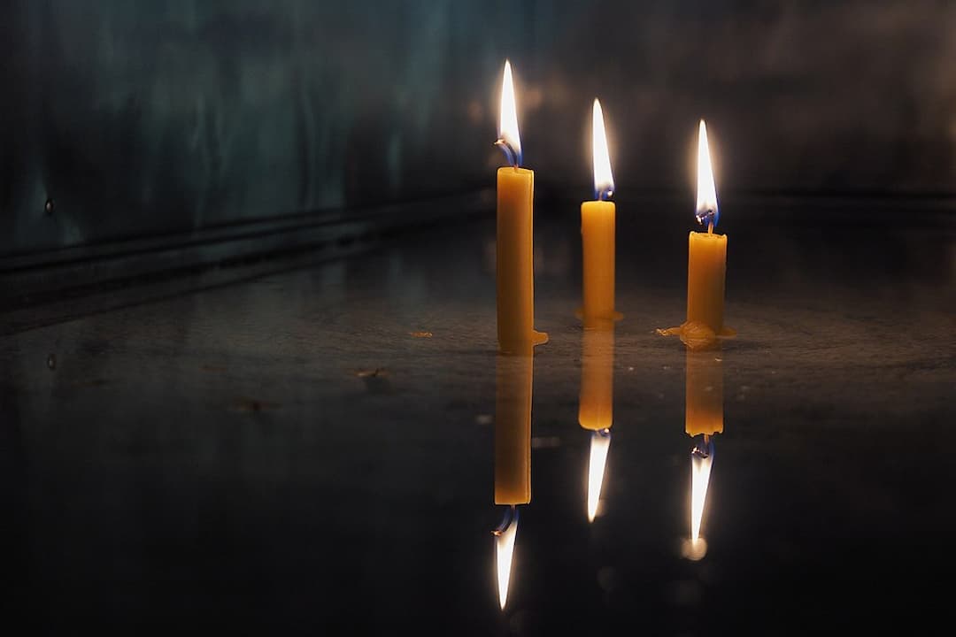 Four lit yellow candles, partially melted, standing upright on a reflective surface with their flames and bodies mirrored below
