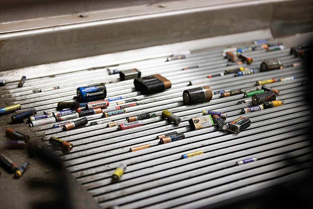 variety of used batteries scattered on a metal conveyor belt, including AA, AAA, C, D, and 9V types, worn labels and corroded surfaces visible